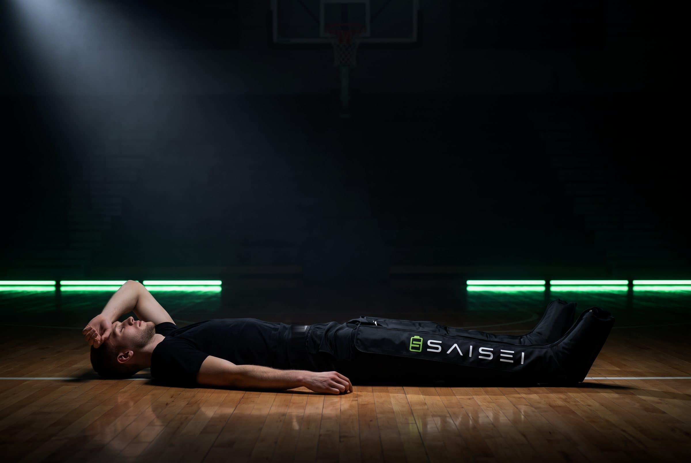 A basketball player lying on the court of a dark arena, legs extended in SAISEI Recovery Boots